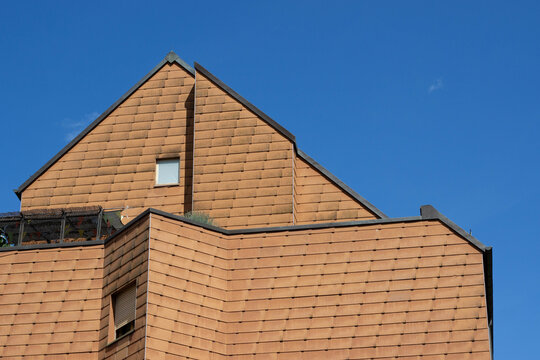 Terracotta tiled house facade with steep gabled roof, small window, angular walls, and clear blue sky in an architectural exterior detail