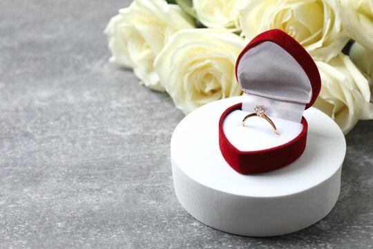 Engagement ring in red box on white podium with roses on grey background, copy space