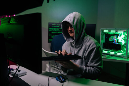 Mysterious anonymous hacker in gray hoodie at desk in dark room, typing on keyboard. Green light from monitors and PC case illuminates him. Cybersecurity and crime concept.