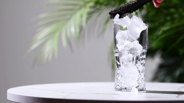 Placing ice in highball glass for cold drink preparation. Tall crystal tumbler with ice cubes on white table. Summer refreshment and hydration lifestyle aesthetic background