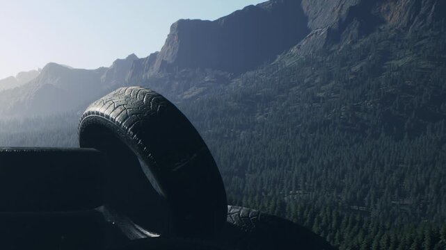 Dark rubber tires sit stacked in the foreground with a vast mountain range and dense evergreen forest covering the background landscape under a clear blue sky during the bright morning time of day.