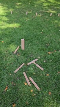 Wooden batons lie crossed on the grass around a single kubb block in a sunny park. Traditional wooden games and unplugged family time for vertical lifestyle content.