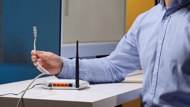 Person holding an unplugged ethernet cable beside a wireless router, preparing a wired internet connection at a desk in a home or office network setup with wi fi access
