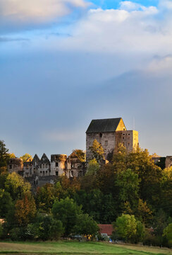 Historic stone castle and ruined medieval buildings above a forested landscape beneath dramatic blue and cloudy skies, European architectural heritage landscape
