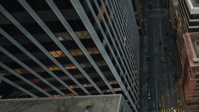 Tall modern buildings stand above an empty city street with parked cars during the night in New York City as the urban landscape remains calm and quiet with warm office lights glowing from the windows