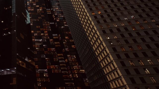 Glowing windows of tall office buildings rise against a dark sky in the heart of New York City during the night as city lights shine from inside many rooms across the tall modern glass structures