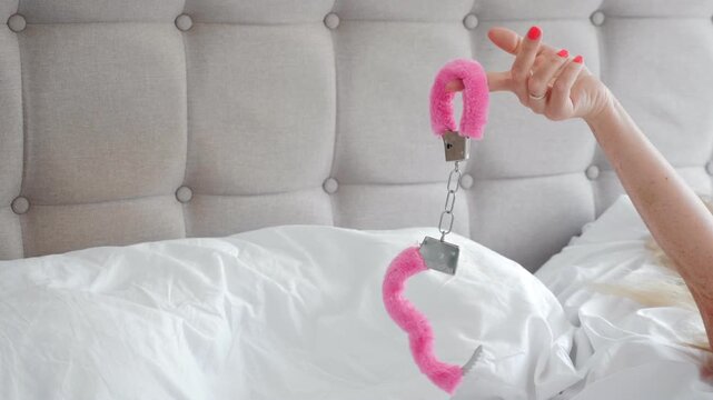Hand holds pink fuzzy handcuffs above rumpled white bedding beside a tufted headboard