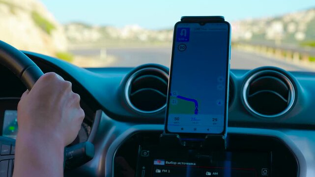 Driver using gps navigation app on smartphone while driving. Driver navigating with a map application on a smartphone mounted on the car dashboard. The vehicle is moving along a sunny road