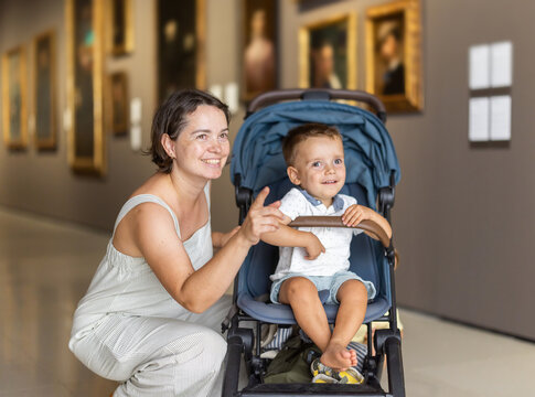 Mother introduces her children to art; woman with toddler in stroller and preschool-age girl are looking at paintings at museum. Culturally and intellectually engaging leisure activity