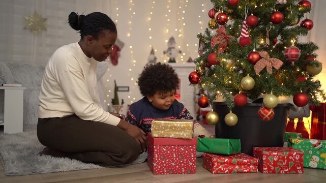 African American mother and her cute son stacking Christmas gifts under a decorated tree at home