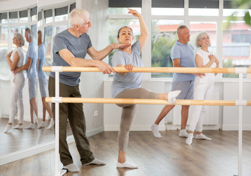 Elderly woman in sportswear, participating in ballet class, mastering developpe movement at barre with aged group and skilled senior male instructor in bright choreography studio