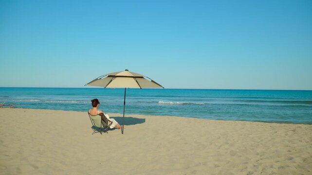 Woman relaxing on sandy beach under umbrella. Woman sitting on a camping chair under a parasol enjoys a peaceful sunny day, looking at the calm sea waves washing ashore on a beautiful sandy beach