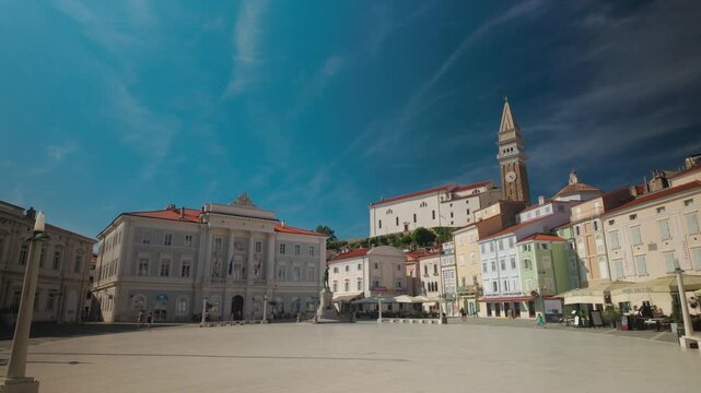Static wide shot of the picturesque Tartini Square in Piran, Slovenia, showcasing historical Venetian architecture and Saint George's Parish Church bell tower under a bright summer sky.