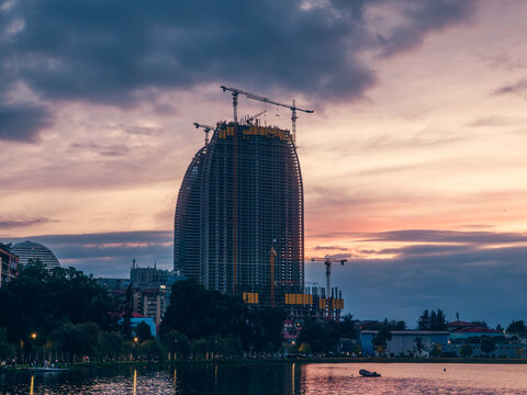 Skyscraper construction and urban development reshape city skyline at sunset as cranes rise above unfinished tower near lake with modern architecture and evening clouds