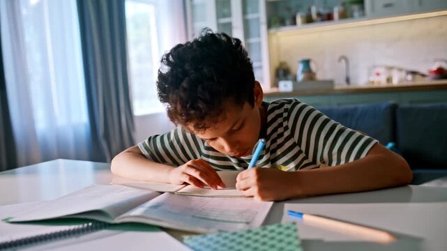 Schoolboy doing homework kitchen table. Mixed race pupil writing exercise book.