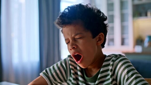 Yawning schoolboy writing homework notebook. Mixed race kid studying home desk.