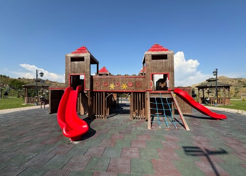 Wooden playground castle with red slides at K&ouml;srelik Pond park in Ankara with black pine (Pinus nigra) and Russian olive (Elaeagnus angustifolia) trees under a clear blue sky on a sunny day.