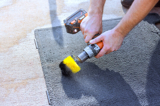 Hands guide drill with yellow brush across gray carpet on concrete surface outdoors.