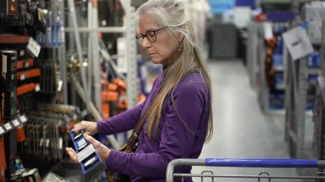 A senior woman examines items in the hardware aisle, exploring tools and home improvement supplies in a large store.