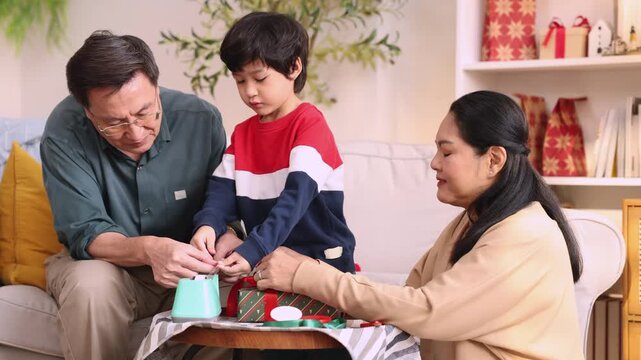 Little boy reaches for adhesive tape while grandparents wrap Christmas gifts together, joining busy hands around patterned paper, enjoying childhood discovery through family cooperation.