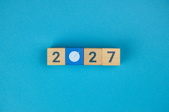 A concept featuring stacked wooden cubes, an arrow icon, and a target. It represents goals, strategy, financial planning, and risk management for the new year, 2027. Light blue background.