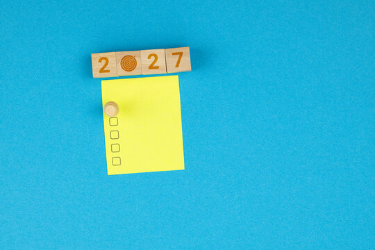 To-do list on a yellow sticky note placed beneath wooden blocks displaying the year 2027. A concept representing goals, organization, and New Year&rsquo;s resolutions, set against a blue background.