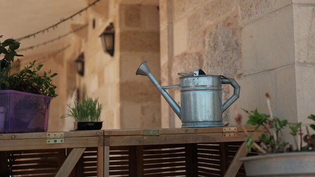 A galvanized metal watering can sitting on a wooden table outdoors next to potted plants