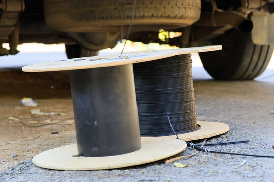 Fiber spool holding black cable beneath parked vehicle, with wooden flanges, asphalt surface, industrial repair setting, and scattered debris creating practical outdoor work scene