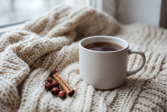 Steaming mug of hot tea on cream chunky knit blanket by a winter window with cinnamon stick and dried rose hips, cozy hygge autumn morning still life