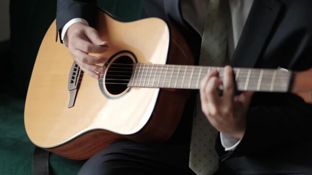 A man dressed in a formal suit and tie sitting on a green sofa playing an acoustic guitar