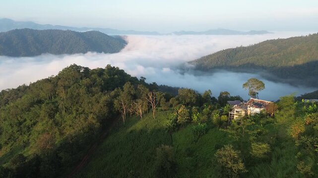Aerial lush green forest mountain landscape with layered ridge, misty valley, quiet lake, rural house, soft cloud sea, tropical vegetation, peaceful panorama beneath pale sunrise sky