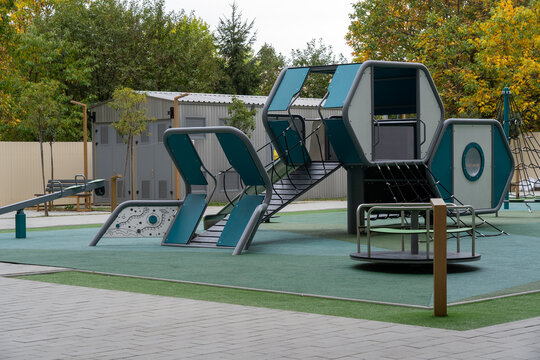 Modern outdoor children's playground with futuristic climbing structures, rope bridges, slides and roundabout on green rubber safety flooring surrounded by autumn trees