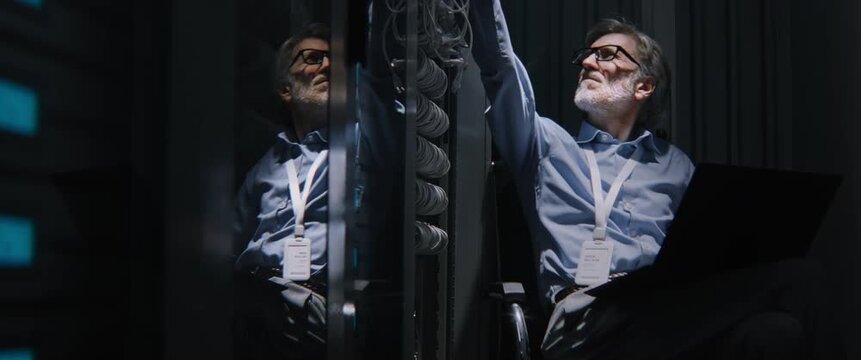 Male Working From Server Aisle with Laptop, Checking Overhead Fiber Lines. Fatigued by Heavy Workload, Man Performs Infrastructure Auditing, Legacy Technology Upgrades, and Connectivity Verification.