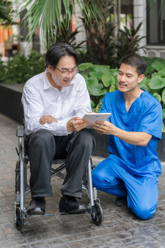 A male Asian doctor in blue scrubs kneels beside a smiling senior patient in a wheelchair outdoor, sharing information on a digital tablet during a consultation.