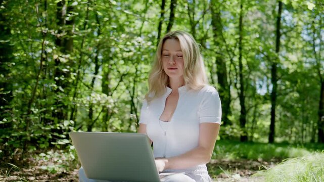 Young woman sits in forest clearing, laptop on lap. She relaxes, eyes closed, smiling softly. Sunlight filters through green trees around her. Ideal for remote work, nature therapy