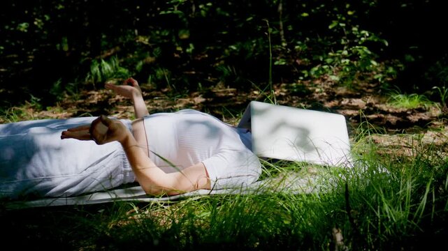 Woman lies on grass, reading a book under trees. She relaxes, surrounded by sunlight filtering through leaves. Her calm expression shows peaceful enjoyment. Perfect for scenes of relaxation