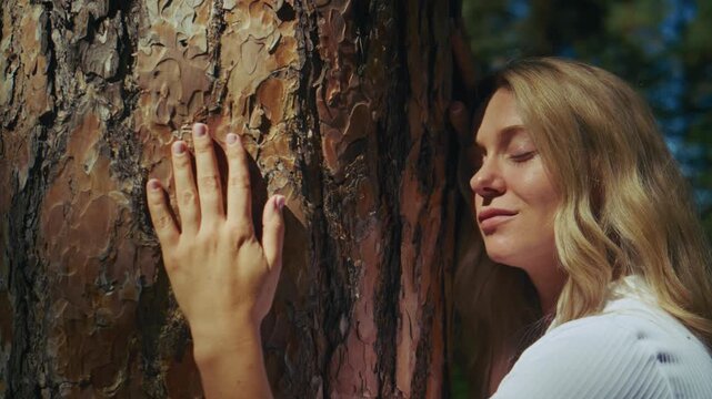 Woman gently touches pine tree bark, eyes closed in calm reflection. Golden sunlight filters through forest canopy above. Scene evokes peace, connection with nature, mindfulness