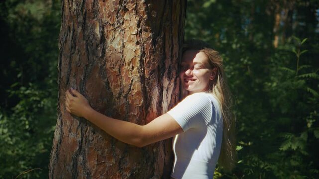 Young woman embraces large pine tree trunk in sunlit forest. Her smile shows peace and connection to nature. Shot captures serene moment of personal reflection outdoors