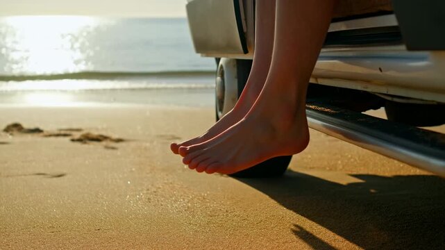 Bare feet sway above beach sand beside open vehicle doorway, with gentle shoreline waves and bright water reflections behind dangling legs in warm sunlight from low side view