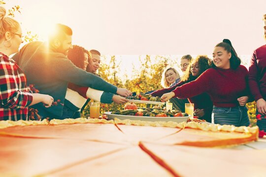 Friends reaching for roasted vegetables at a long outdoor autumn harvest table with giant pie in warm golden sunlight