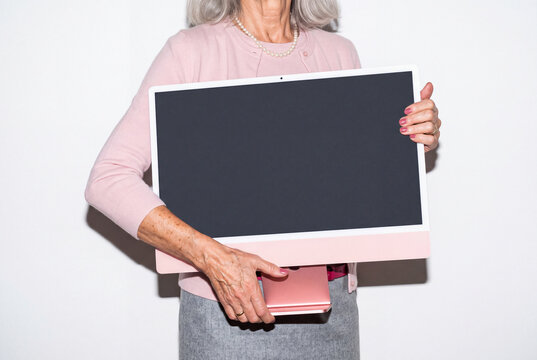 A senior woman with grey hair, a pearl necklace and blush cardigan hugs a pink all-in-one desktop computer against her hip. Direct flash shoot.