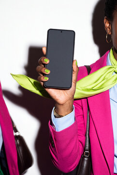 A woman in a magenta blazer and lime silk neckerchief raises a smartphone with nails painted to match, a gold hoop earring and watch catching the light.