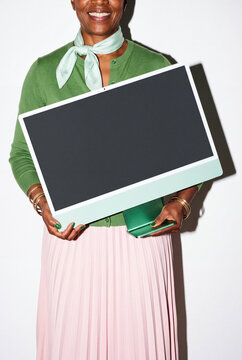 A senior woman in a green cardigan, mint silk scarf and pink pleated skirt smiles broadly, clearly pleased with the new all-in-one computer she holds. Gold bangles stack on both wrists. 