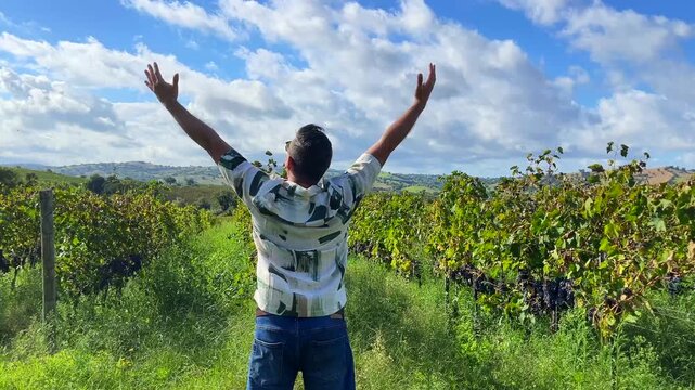 Latin backpacker stylish tourist male with arms outstretched embracing the beautiful Italian countryside landscape while standing in a vineyard under a clear blue sky, experiencing joy and relaxation