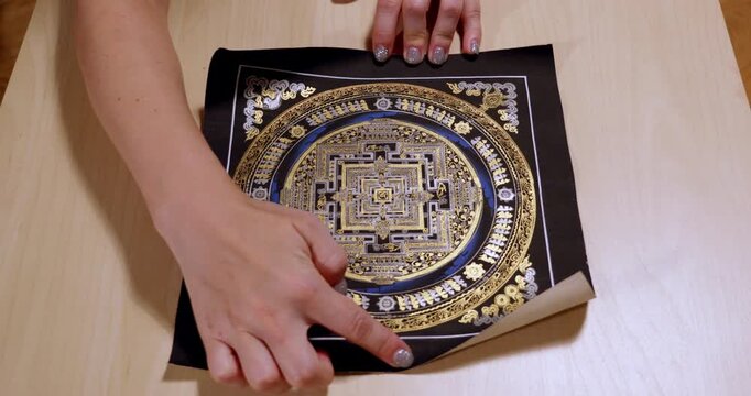 Traditional Nepalese mandala scroll opens under hands revealing golden squares on black inside blue circular bands and Buddhist symbols before fingers roll the painting closed on a wooden table