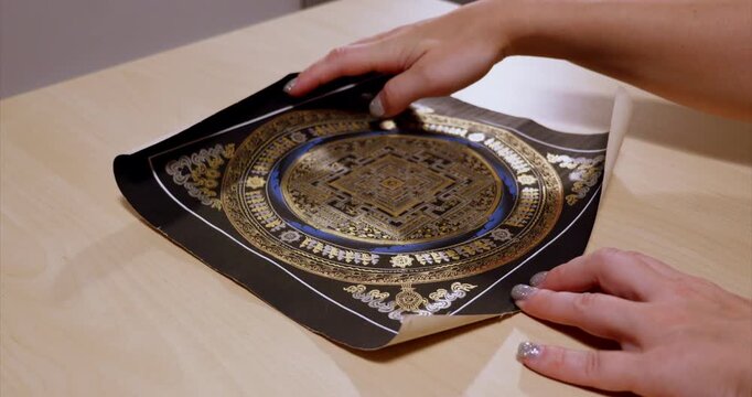 Nepalese Buddhist mandala on black is unrolled by hands on a wooden table exposing a golden square center encircled by blue bands and symbol rows while the corners of the painting curl up