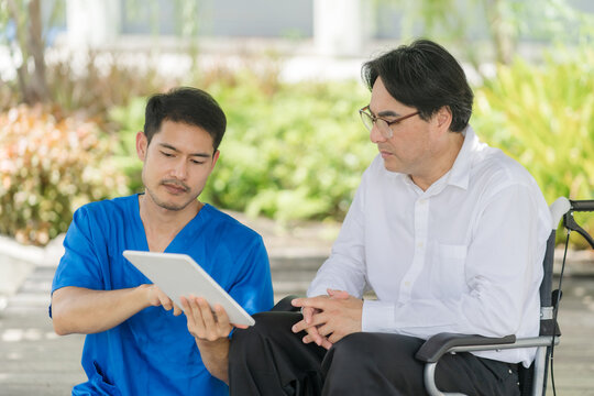 A male Asian doctor in blue scrubs sits with a senior patient in a wheelchair outdoor, showing medical results on a digital tablet during a consultation.