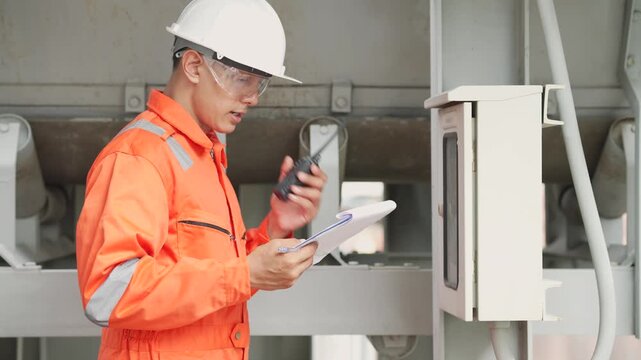 Male industrial engineer checks electrical control panel while reviewing maintenance documents on clipboard at industrial plant. Technical inspection, workplace safety.
