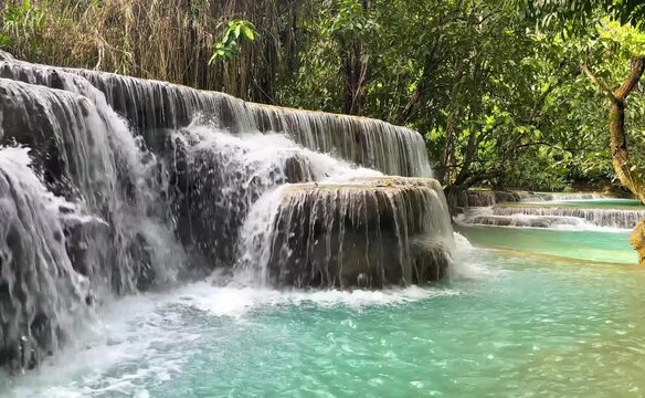 Curtains of water spill over rounded rock ledges into a turquoise pool at Kuang Si Waterfall near Luang Prabang, Laos, surrounded by tropical forest.