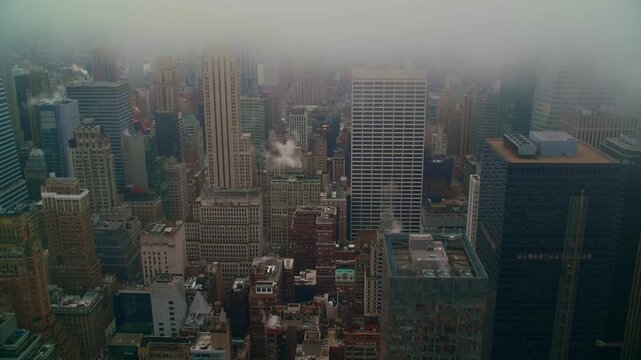 Closely packed Manhattan buildings extend beneath a low cloud layer, with rooftops and narrow streets creating a dense pattern in the elevated city view. A winter view of New York City, United States.
